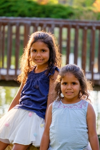 Two young girls with curly hair sitting on a huge rock outdoors looking at camera together - Australian Stock Image