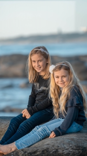 Under 10 girls sitting on rock at beach - Australian Stock Image