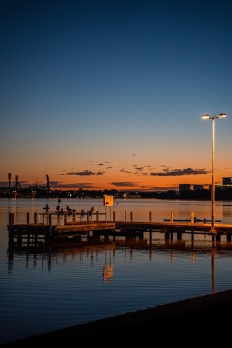 vertical shot of a pier during sunset with lamp post, buildings reflected by the water - Australian Stock Image
