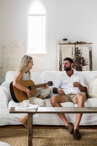 Vertical shot of a woman and a man on a white couch playing guitar and drinking coffee