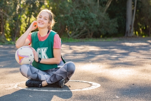 horizontal shot of a young woman sitting on the ground touching her face and the net ball on her lap - Australian Stock Image