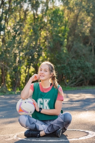 vertical shot of a young woman sitting on the ground touching her face and the net ball on her lap - Australian Stock Image