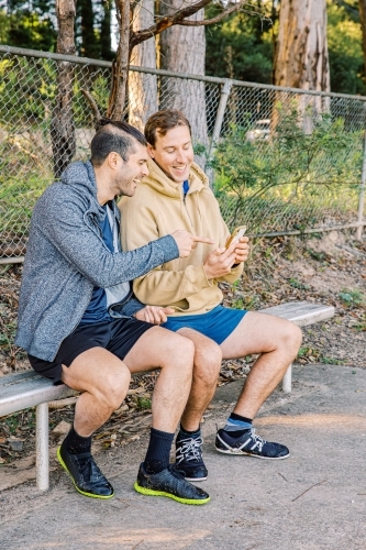 vertical shot of two men sitting on a bench smiling while looking at a cellphone - Australian Stock Image