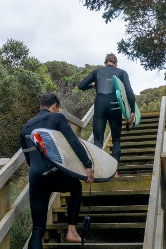 Vertical shot of two men walking up wooden stairs wearing wetsuits and carrying surfboards. - Australian Stock Image