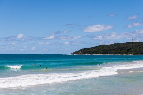 View across the water to Cape Byron and lighthouse from  Byron Bay with surfers - Australian Stock Image