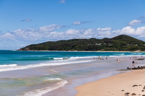 View of Cape Byron and lighthouse from the beach at Byron Bay with surfers, rocks, sandy foreshore - Australian Stock Image