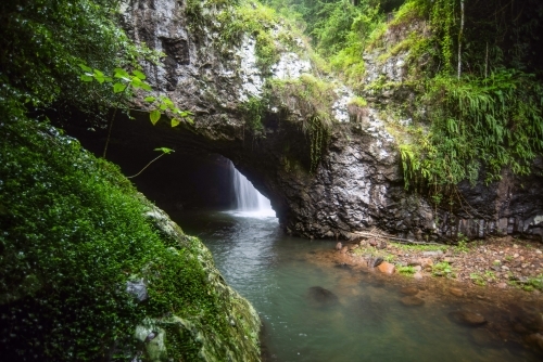 View of the waterfall through an opening of a cave mouth with the greenery surrounds - Australian Stock Image