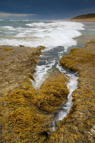 Waves rushing into a rock pool covered in seaweed under a dark sky. - Australian Stock Image