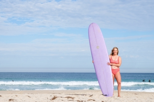 Wide view of woman surfer standing on beach in bikini holding a longboard surfboard - Australian Stock Image