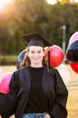 Woman holding balloons in park smiling wearing graduation cap and gown - Australian Stock Image