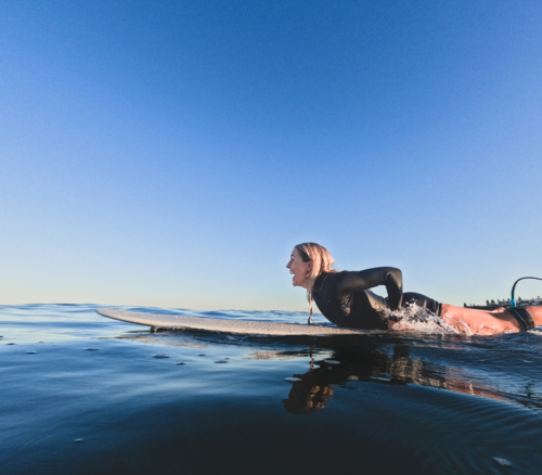 Woman in summer wetsuit lying on longboard surfboard paddling looking out to glassy sea laughing - Australian Stock Image