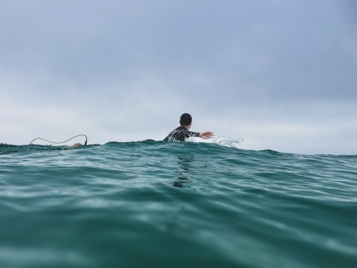 Woman looking away paddling surfboard across ocean taken at water level with overcast sky - Australian Stock Image