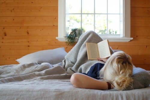 Woman lying in bed reading book with timber walls and window in background - Australian Stock Image