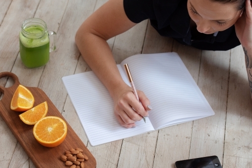 woman lying on floor journalling with green juice oranges almond healthy snack and phone - Australian Stock Image