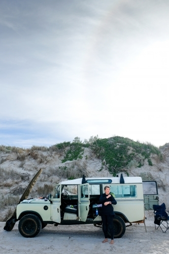 Woman putting on wetsuit on beach in front of vintage Land Rover with surfboard leaning on car - Australian Stock Image