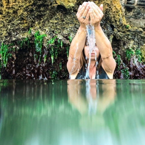 Woman sitting in glassy ocean and cupping water with coastal limestone rocks in background - Australian Stock Image
