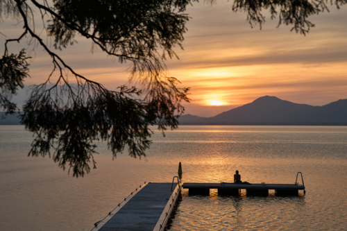 woman sitting on jetty watching sunset with tree in foreground - Australian Stock Image