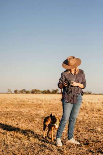 Woman standing in a dusty and dry grassy farm paddock holding a camera - Australian Stock Image