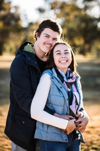 Woman standing laughing in front of boyfriend arms wrapped around her holding hands - Australian Stock Image