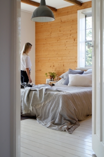 Woman walking into bedroom with grey bed linen, timber wall and window - Australian Stock Image