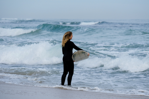 Woman wearing steamer wetsuit holding surfboard looking out at surf - Australian Stock Image