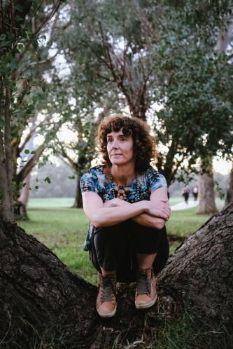 woman with curly hair and casually dressed sitting on tree trunk - Australian Stock Image