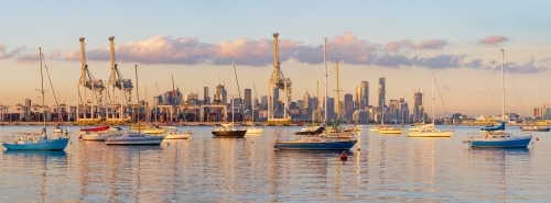 Yachts anchored on still bay in front of large gantry cranes at a cargo dock - Australian Stock Image