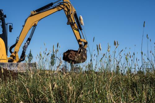 Yellow excavator carrying bucket of dirt across grassy paddock field - Australian Stock Image