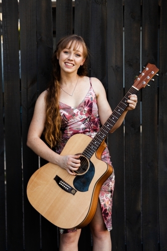 Young adult guitar player playing instrument outside leaning on black wooden fence - Australian Stock Image