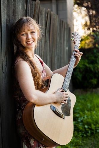 Young adult guitar player playing instrument outside leaning on black wooden fence - Australian Stock Image