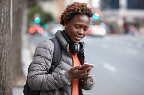 Young African woman text messaging wearing wireless headphones in city - Australian Stock Image