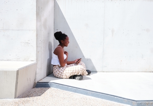 Young African woman using a phone sitting against a concrete wall