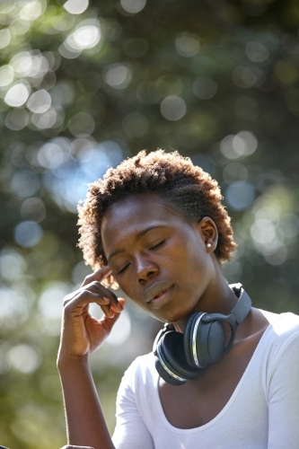 Young African woman with eyes closed wearing wireless headphones at park - Australian Stock Image