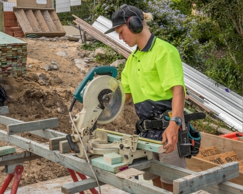 Young apprentice builder measuring and cutting timber with drop saw on residential construction site