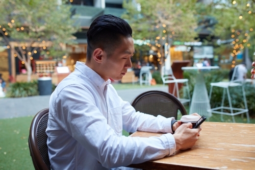 Young Asian man having coffee and checking phone outside at cafe - Australian Stock Image
