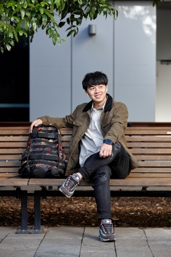 Young Asian man smiling sitting on park bench whilst listening to wireless headphones - Australian Stock Image