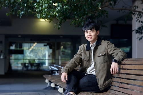 Young Asian man smiling sitting on wooden bench - Australian Stock Image