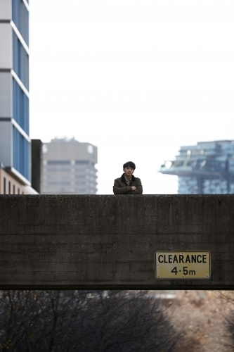 Young Asian man standing on bridge with city in background - Australian Stock Image
