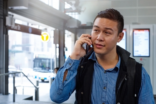 Young Asian man talking on mobile phone at train station entrance - Australian Stock Image