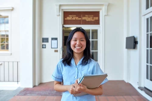Young Asian student in front of school building holding books - Australian Stock Image