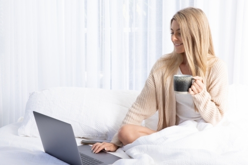 Young blonde lady typing on a laptop in white bed - Australian Stock Image