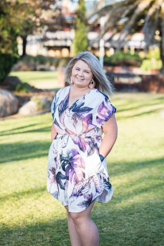 Young blonde woman in early 20s standing in garden with hands in pockets smiling - Australian Stock Image