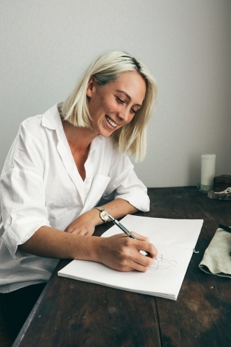 Young blonde woman laughing as she sits at a desk drawing in a sketch book