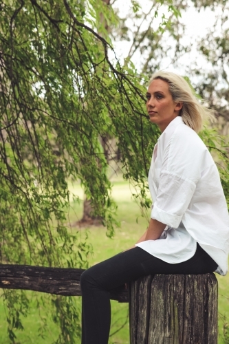 Young blonde woman looking away, sitting on a fence post as wind blows her hair - Australian Stock Image