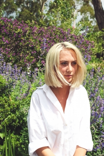 Young blonde woman sits against background of lavender and purple shrub - Australian Stock Image