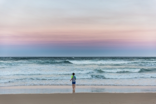 Young boy alone on beach at sunset looking at waves
