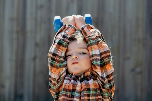 Young boy at home in his backyard lifting a dumbbell weight above his head - Australian Stock Image