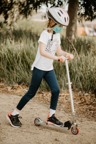 Young boy during the corona virus COVID-19 pandemic wearing a fabric mask and riding scooter - Australian Stock Image