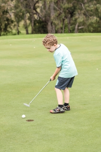 Young boy holding golf club putter hitting ball into hole on green - Australian Stock Image