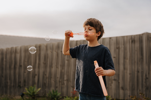 Young boy in black shirt blowing bubbles in their yard. - Australian Stock Image